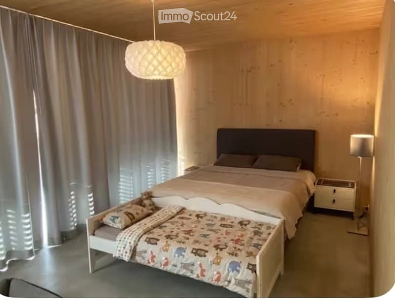 Cozy bedroom with a large bed, nightstand, lamp, and a small child's bed with a colorful blanket and stuffed animal. Wooden walls and a hanging light fixture.