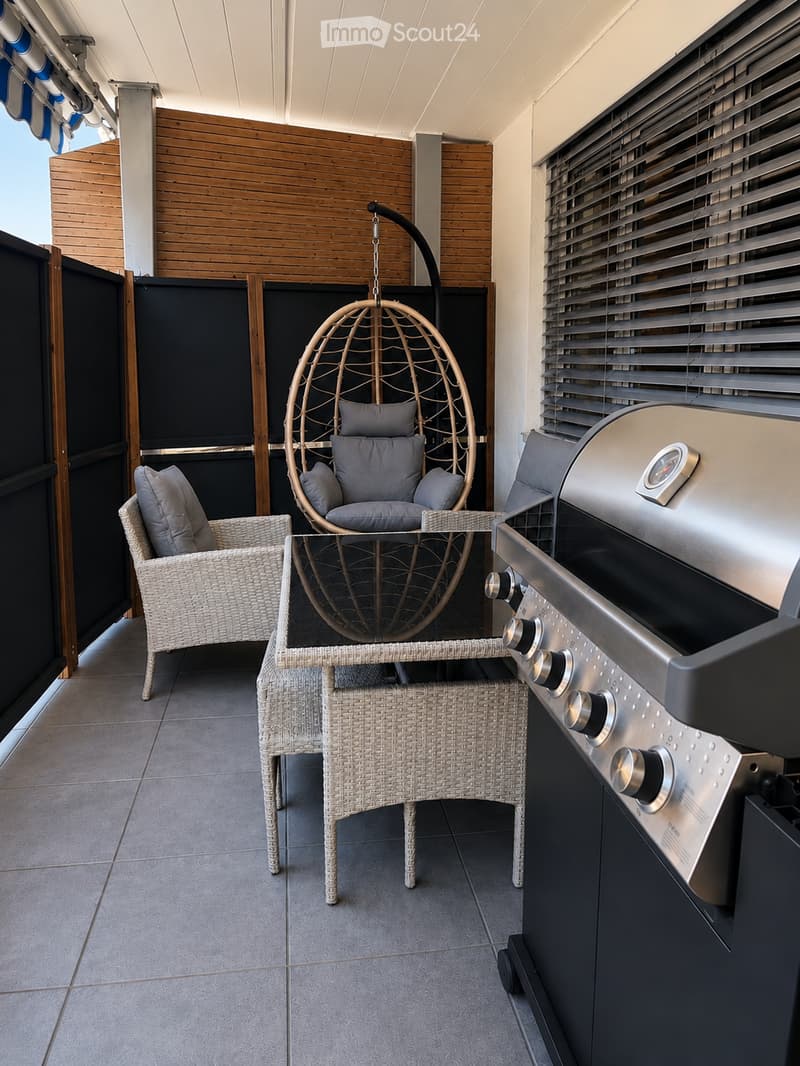 Balcony with a wicker hanging chair, a wicker chair, a small table, and a grill. The space features wooden paneling and a covered ceiling.