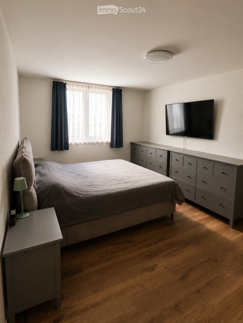 Modern bedroom with a large bed, nightstand, lamp, drawer unit, and wall-mounted television. The room features wooden flooring and large windows with dark curtains.