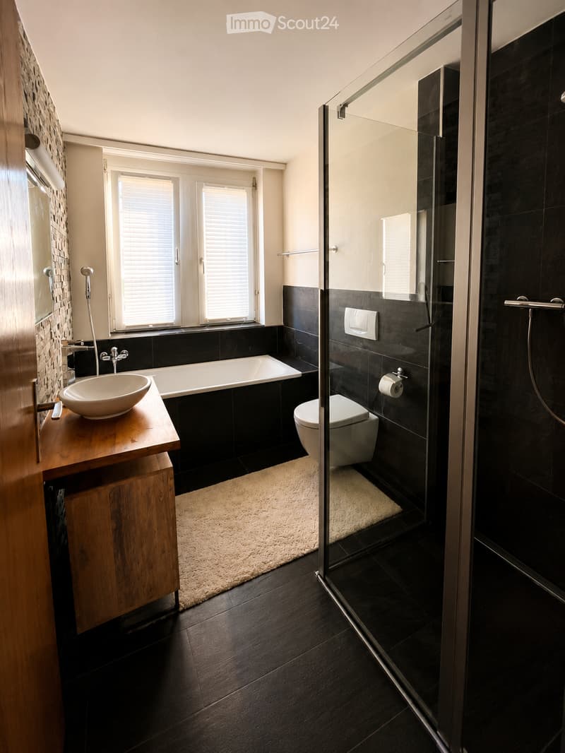 Modern bathroom with dark tiles, a wooden vanity, a bathtub, a toilet, a glass-enclosed shower, and a large window with blinds.