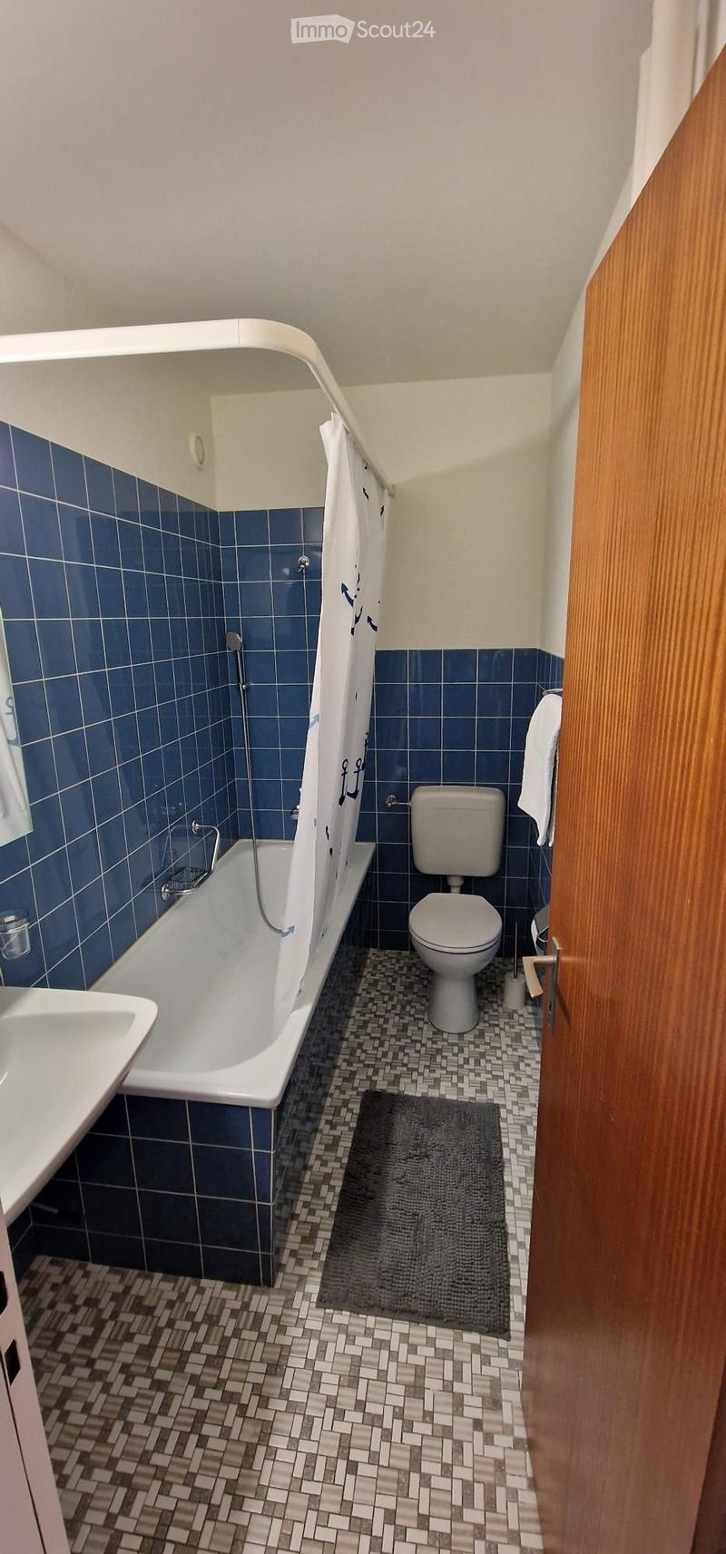 Bathroom with blue tiled walls, a bathtub, toilet, sink, and a shower curtain. The floor has a patterned tile design, and a small rug is placed in front of the bathtub.