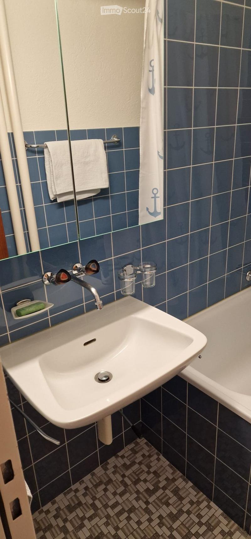 Bathroom with a white sink, chrome faucet, mirror, towel, and a bathtub. Blue tiles on walls and a patterned tile floor.