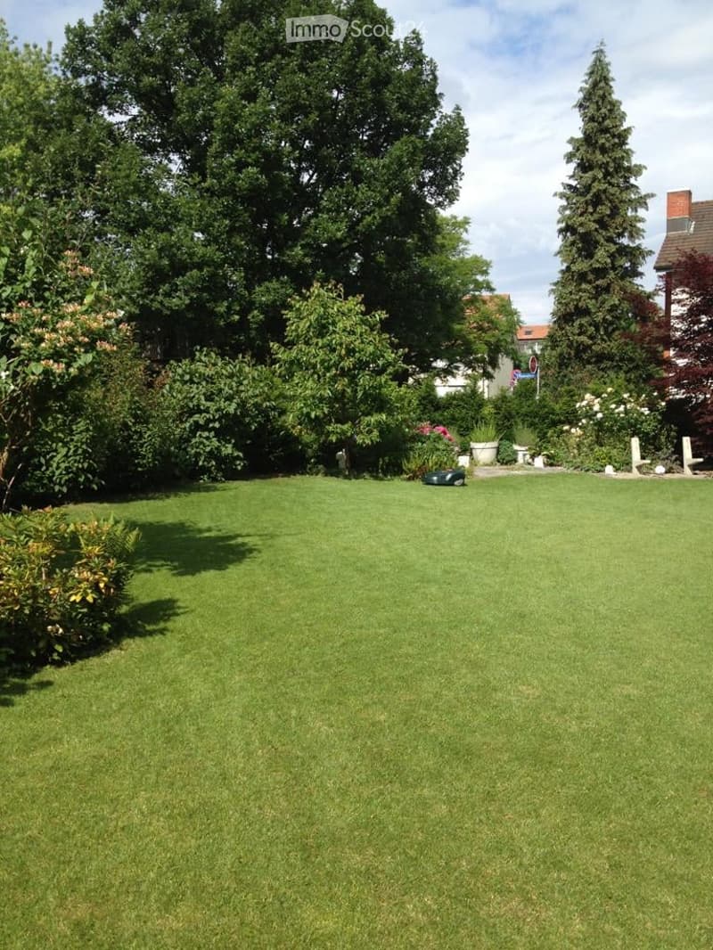 Well-maintained garden with a large, neatly trimmed lawn, surrounded by various trees and shrubs, with a few decorative elements and a small vehicle in the background.
