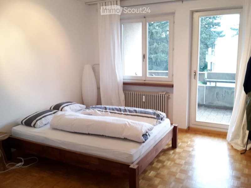 A small, well-lit bedroom with a wooden bed, white bedding, and a radiator under the window. The room features wooden flooring and a sliding glass door leading to a balcony.