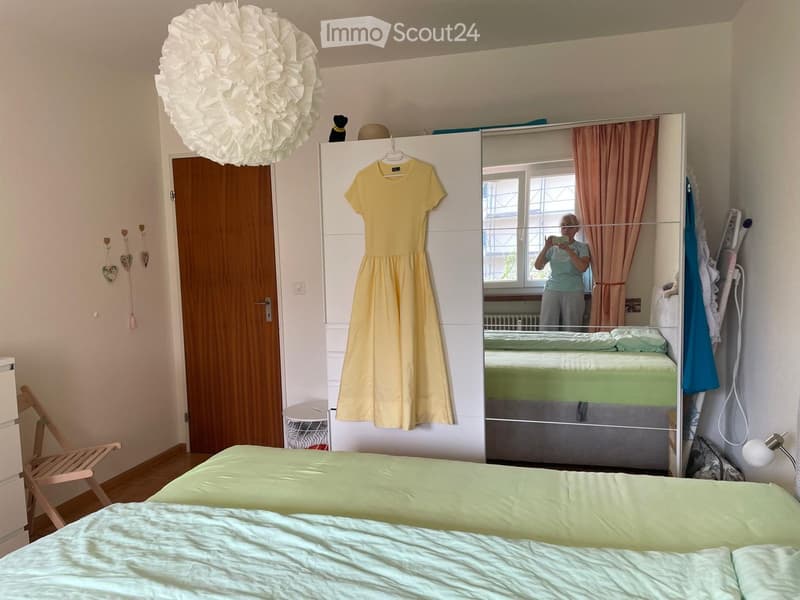 Cozy bedroom with a neatly made bed, a large wardrobe with a hanging dress, a wooden chair, and a round decorative light fixture on the ceiling.