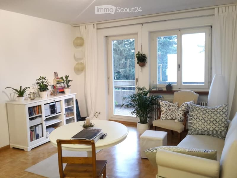 Bright living room with large windows and a sliding glass door leading to a balcony. Features a cream-colored sofa with patterned cushions, a round coffee table, a wooden chair, a white bookcase, and several potted plants.