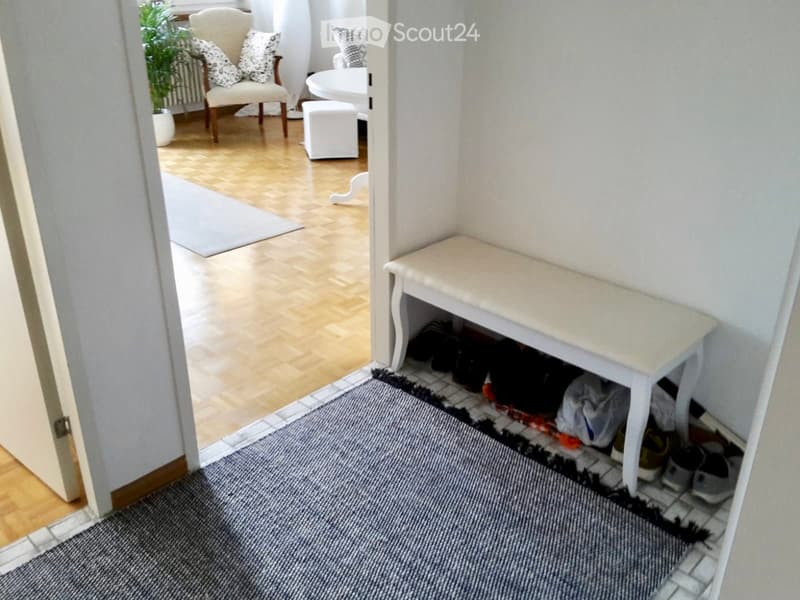 Hallway with wooden flooring, a small white bench, and a gray rug leading to a living area with a chair, small table, and plant.