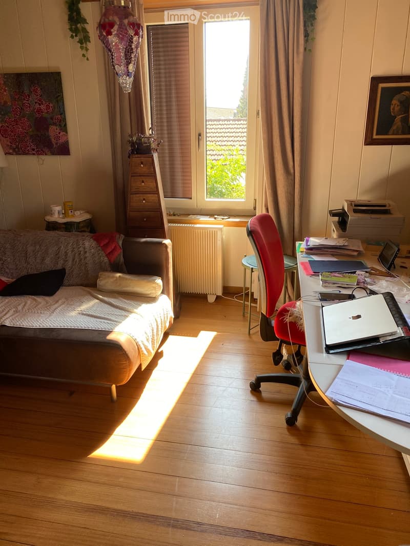A cozy living room with a wooden floor, a sofa with a blanket, a red office chair, a desk with a laptop, and a printer. There is a window with curtains and a radiator below it.