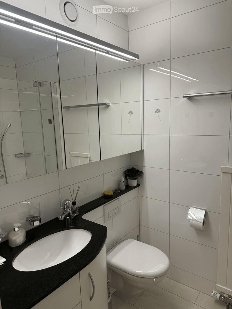 Modern bathroom with white tiles, a black countertop, a white sink, a toilet, a mirror, and a shower area.