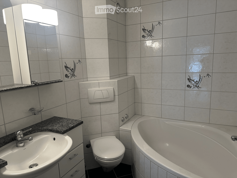 Modern bathroom with a white tiled wall, a bathtub, a toilet, a sink with a granite countertop, and a mirror with a built-in light fixture.