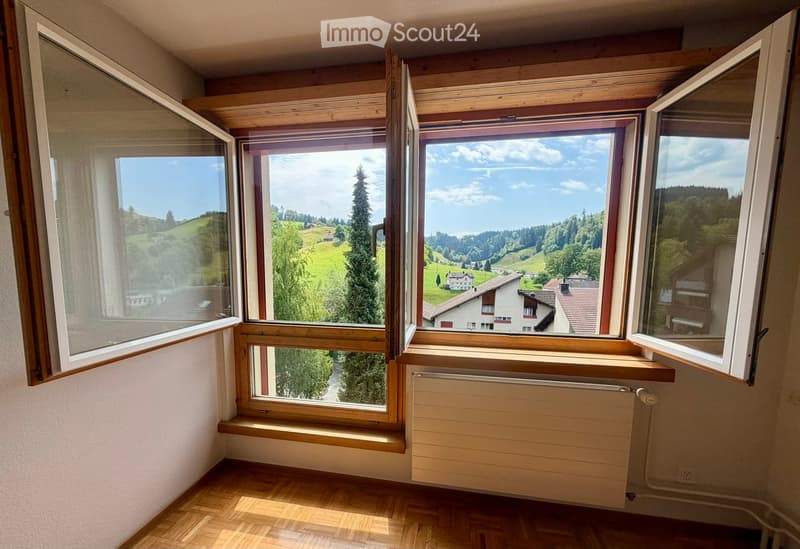 Empty room with wooden flooring, large windows offering scenic views of green hills and houses, radiator, and wooden window sills.