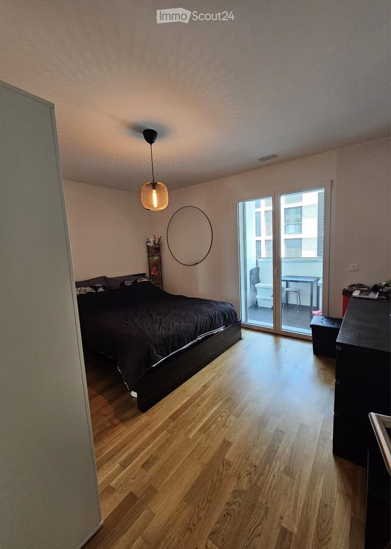 Modern bedroom with wooden flooring, large bed, nightstand, and a balcony with a small table and stool.