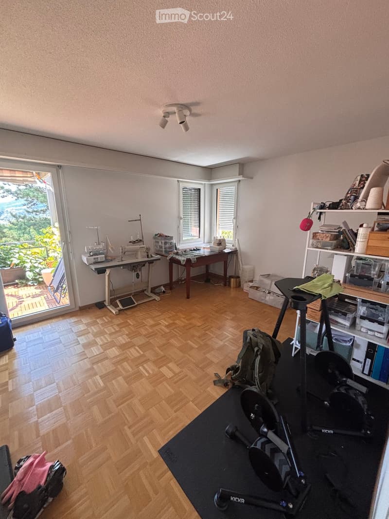 Well-lit room with wooden flooring, large windows, sewing machine, exercise bike, shelves, and various storage items.