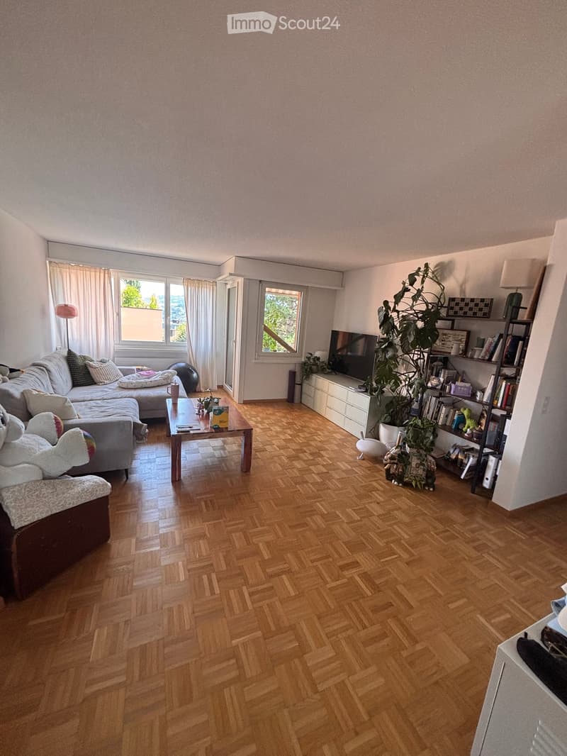 Living room with herringbone wooden flooring, large windows, grey sectional sofa, coffee table, TV, bookshelf, and decorative plants.