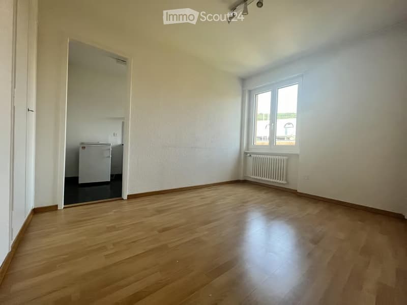 Empty room with wooden flooring, white walls, and a window with a radiator below. A small refrigerator is visible in an adjacent room through an open doorway.
