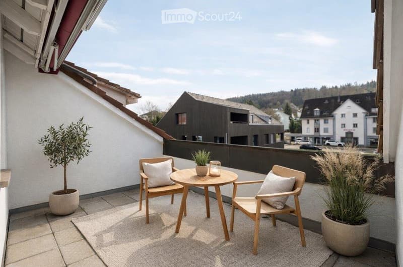 A modern balcony with a small round table, two wooden chairs, and potted plants, overlooking a town with houses and greenery.