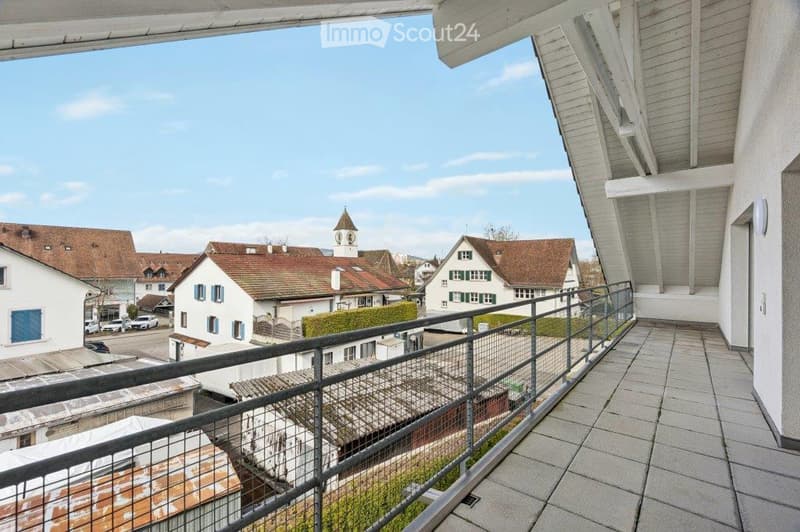 Balcony with metal railing overlooking a town with traditional houses, a church tower, and a clear blue sky.