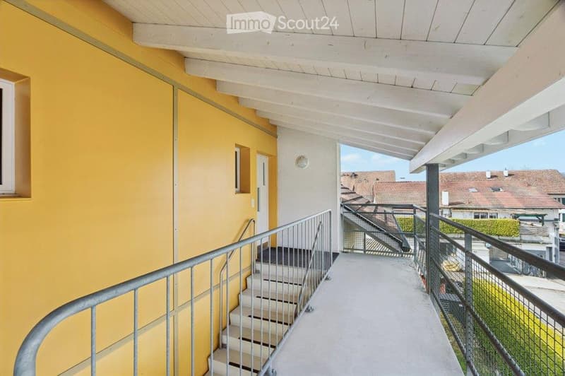 Balcony with white wooden ceiling, metal railing, and steps leading to a lower level, overlooking a residential area with visible rooftops.