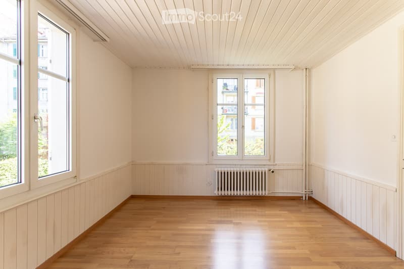 Empty room with wooden flooring, white walls, and large windows offering a view of the outside. A radiator is visible on one wall.
