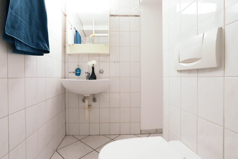 Clean, modern bathroom with white tiles, a white sink, a mirror with a built-in shelf, a toilet, a towel, and a small flower vase on the sink counter.