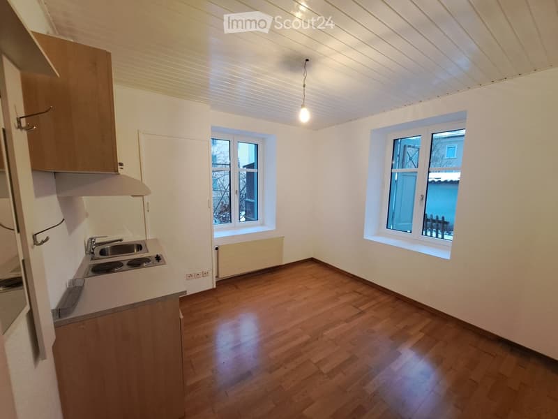 Empty kitchen with wooden flooring, white walls, and two windows. Features a countertop with a sink and electric stove, and a wooden cabinet with a range hood above.