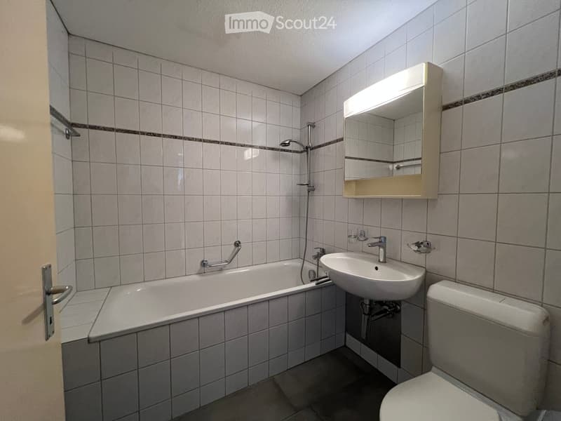 Bathroom with a bathtub, sink, toilet, mirror, and showerhead. The walls are tiled with a decorative border, and the floor is tiled.