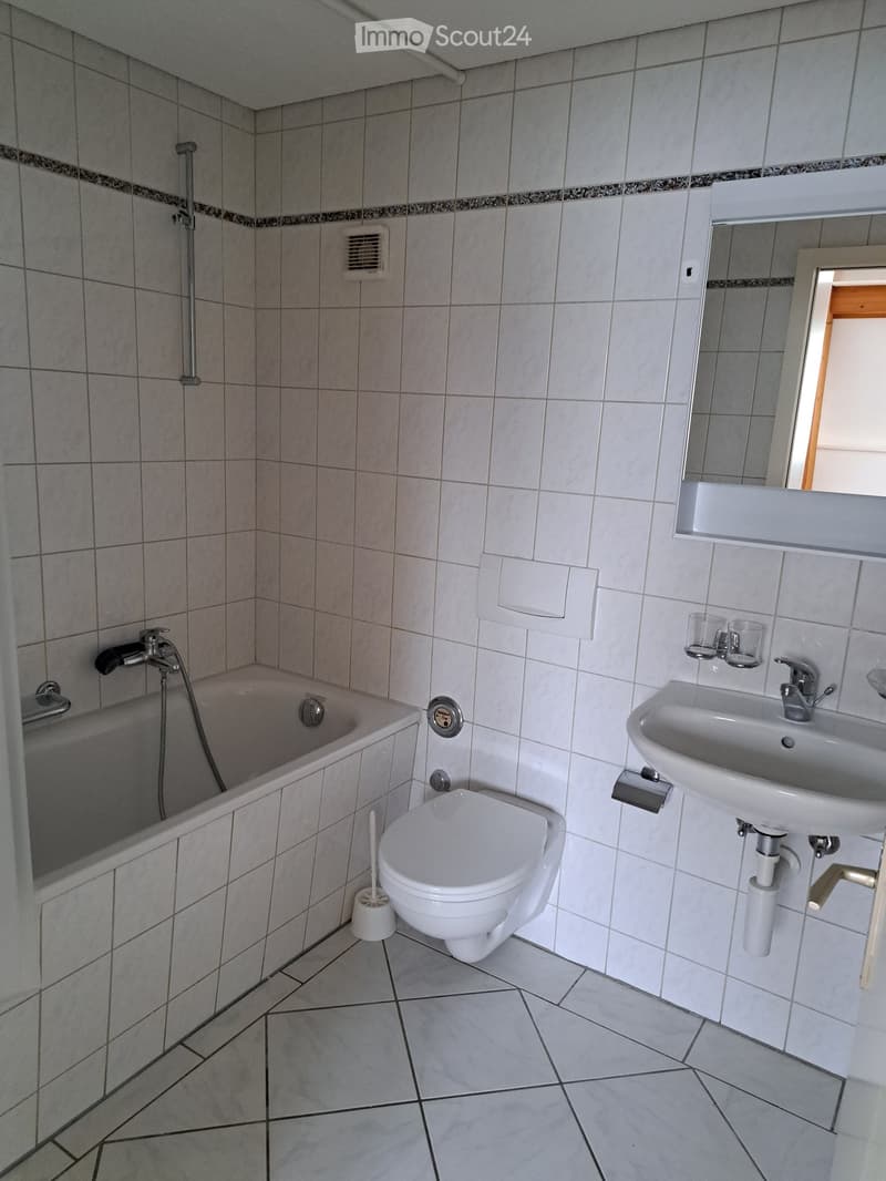 Modern bathroom with a bathtub, toilet, sink, and mirror. The walls and floor are tiled in light gray with a decorative border. A small window is visible on the right side.