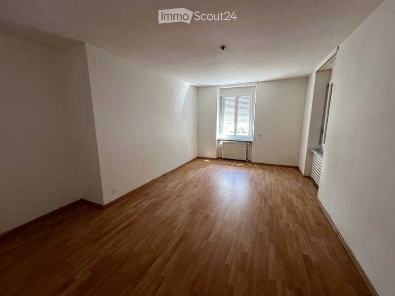 Empty room with wooden flooring, white walls, and a window with blinds. Radiators are visible along the walls.