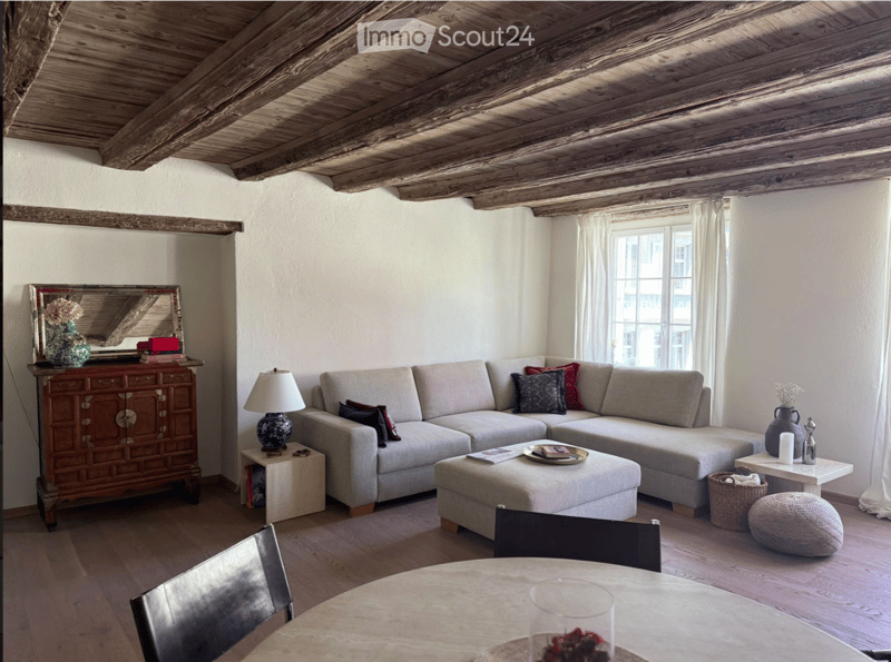 Living room with exposed wooden ceiling beams, grey sectional sofa, ottoman, small table, and various decorative items including a mirror, vase, and lamp.