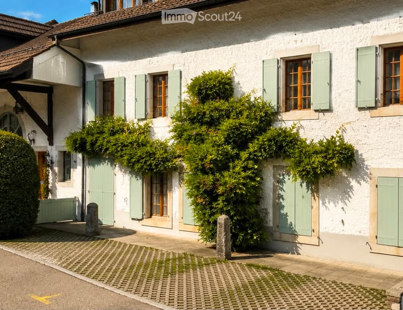 A charming white house with green shutters and a well-maintained garden, featuring neatly trimmed bushes and a cobblestone pathway.