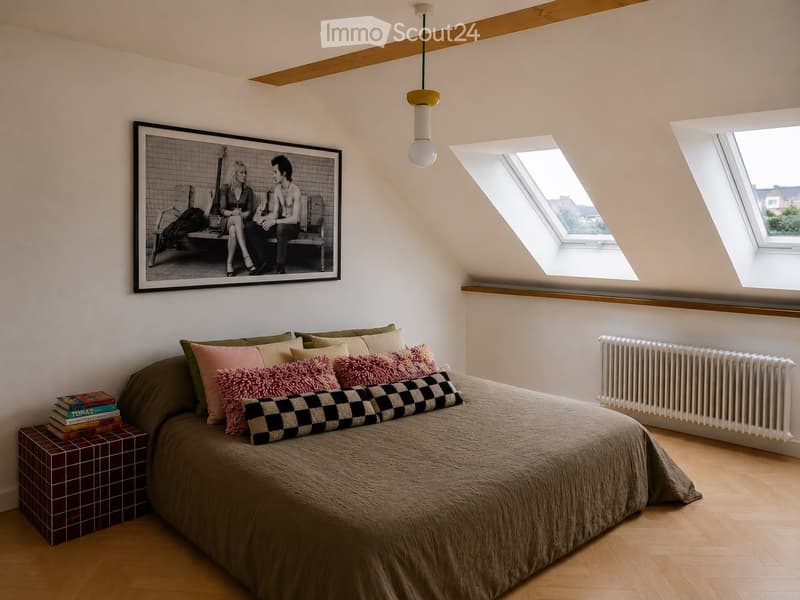 A cozy attic bedroom with wooden floors, slanted ceilings, and skylights. Features a large bed with multiple pillows, a stack of books on a small table, a radiator, and a large framed picture on the wall.