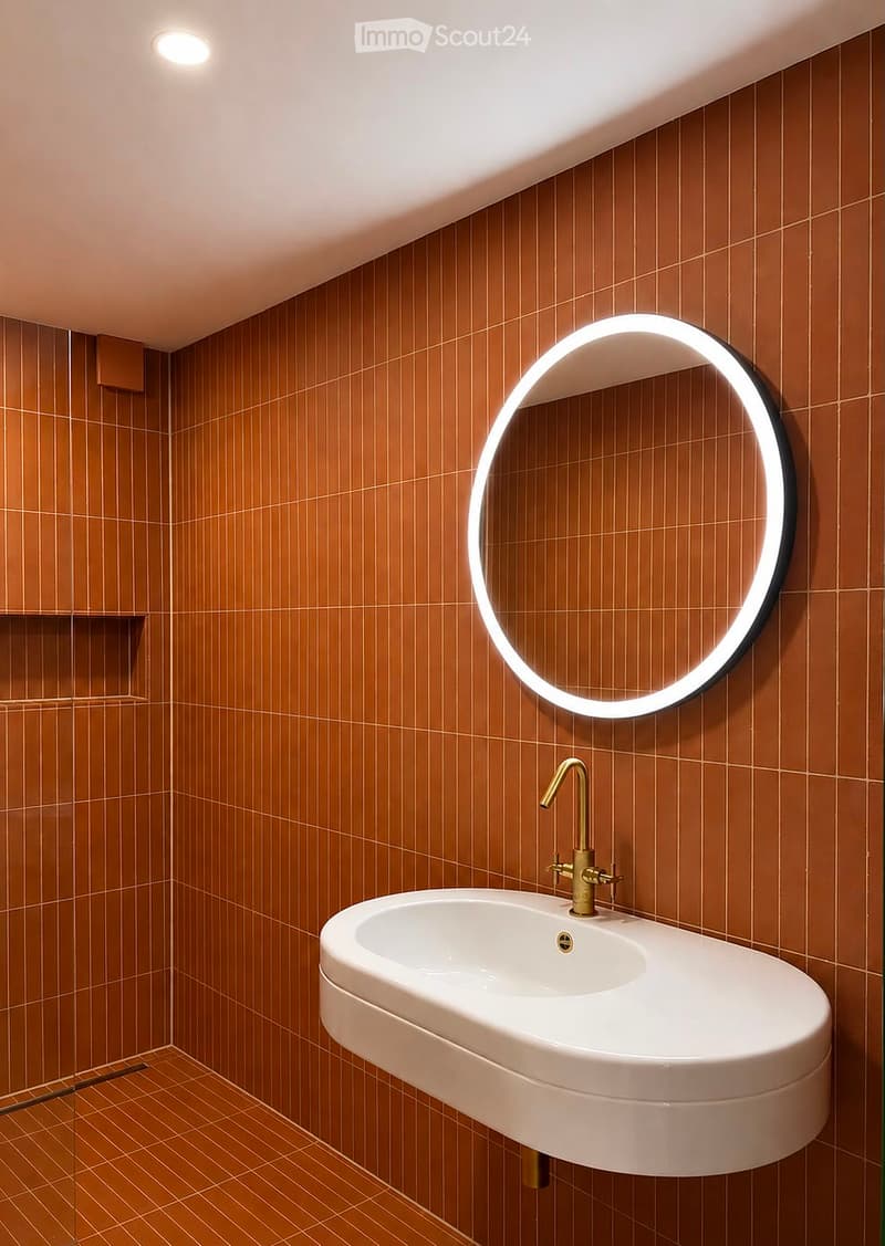 Modern bathroom with brown tiled walls and floor, white sink with gold faucet, and a round illuminated mirror above the sink.
