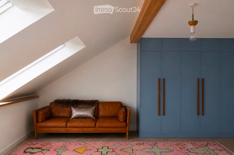A cozy attic living space with a brown leather sofa, a decorative pillow, a colorful rug, and built-in blue cabinets.