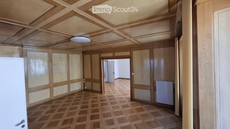 Empty hallway with wooden paneled walls, a patterned wooden floor, and a white ceiling with recessed lighting. There are doors on both sides and a radiator on the right wall.