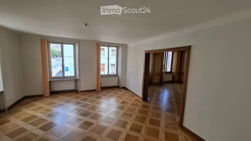 Empty room with wooden flooring, large windows, and a door leading to another room. No furniture or equipment is present.