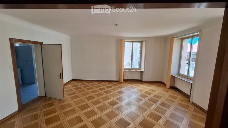 Empty room with wooden parquet flooring, white walls, and large windows. There is a door leading to another room and radiators under the windows.