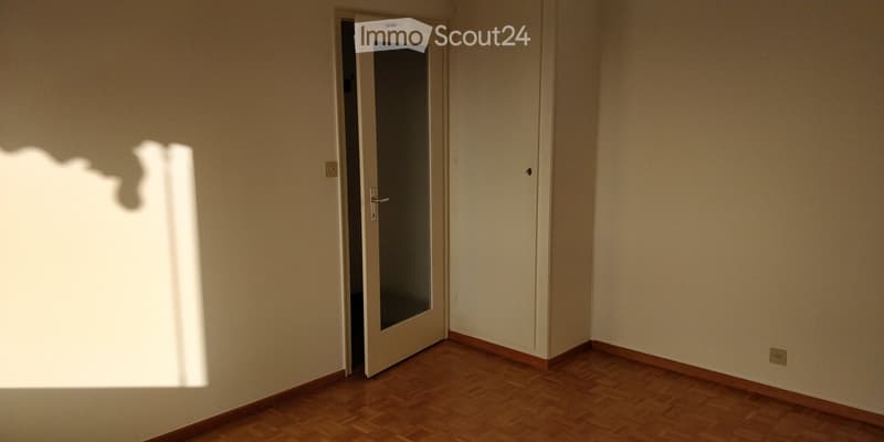 Empty room with wooden flooring and white walls, featuring a partially open door with a glass panel leading to another space.