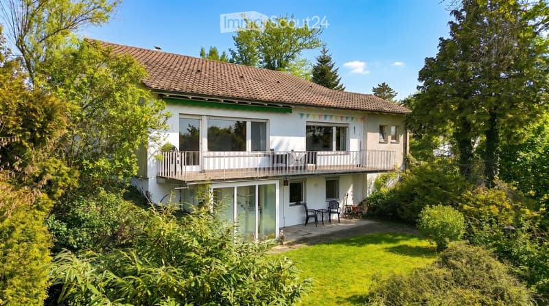 Two-story house with a tiled roof, white exterior, large windows, and a balcony on the upper level. The house is surrounded by greenery and has a small patio area with outdoor furniture.