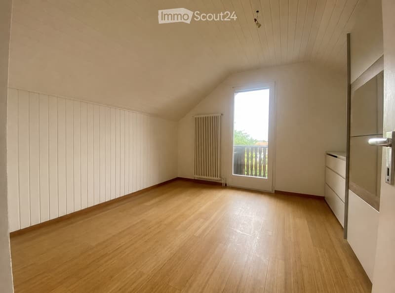 Empty room with wooden flooring, white walls, and a large window with a view of a balcony. The room features a radiator and a door with a modern handle.
