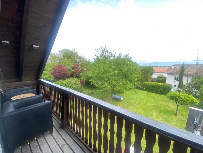 Balcony with wooden decking, black leather sofa, small round table, overlooking a green garden and distant hills.