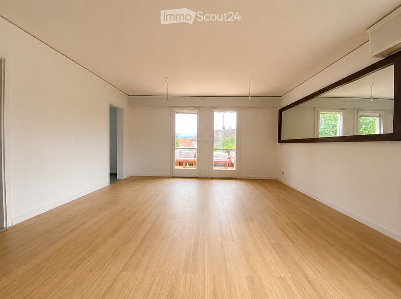 Empty room with light wooden flooring, white walls, large sliding glass doors leading to a balcony, and a large mirror on one wall.