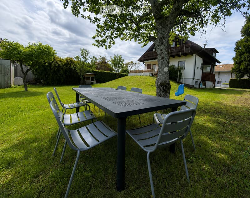 A grassy outdoor area with a metal table and chairs, a swing hanging from a tree, and a house with a balcony in the background.