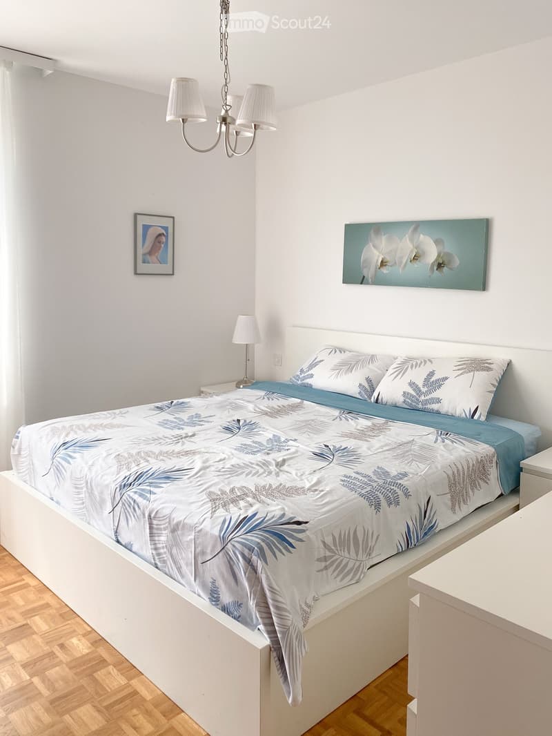 A modern bedroom with a white bed, blue and white bedding, a nightstand with a lamp, and artwork on the walls.