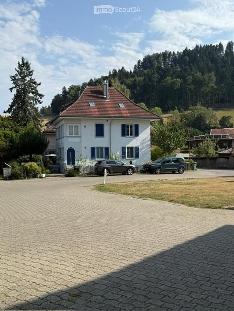 Two-story white house with blue shutters and a red-tiled roof, surrounded by trees and a parking area with two cars.