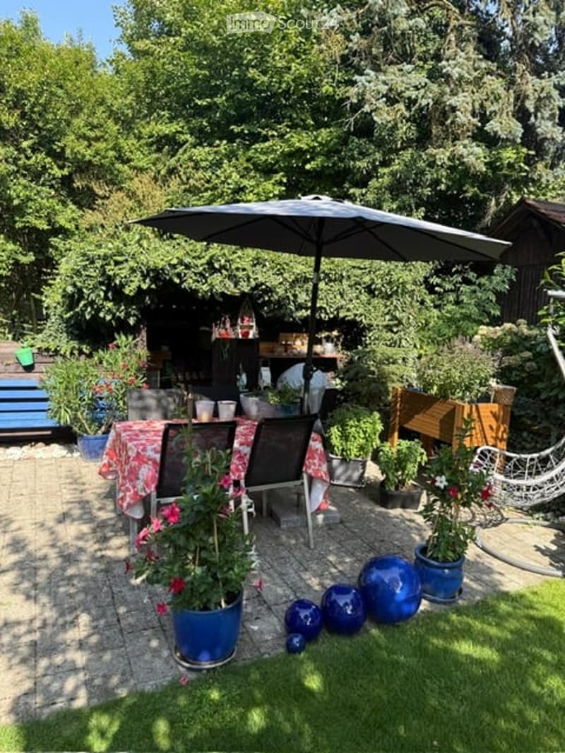 A well-maintained garden patio with a dining table, chairs, umbrella, potted plants, and decorative blue spheres.