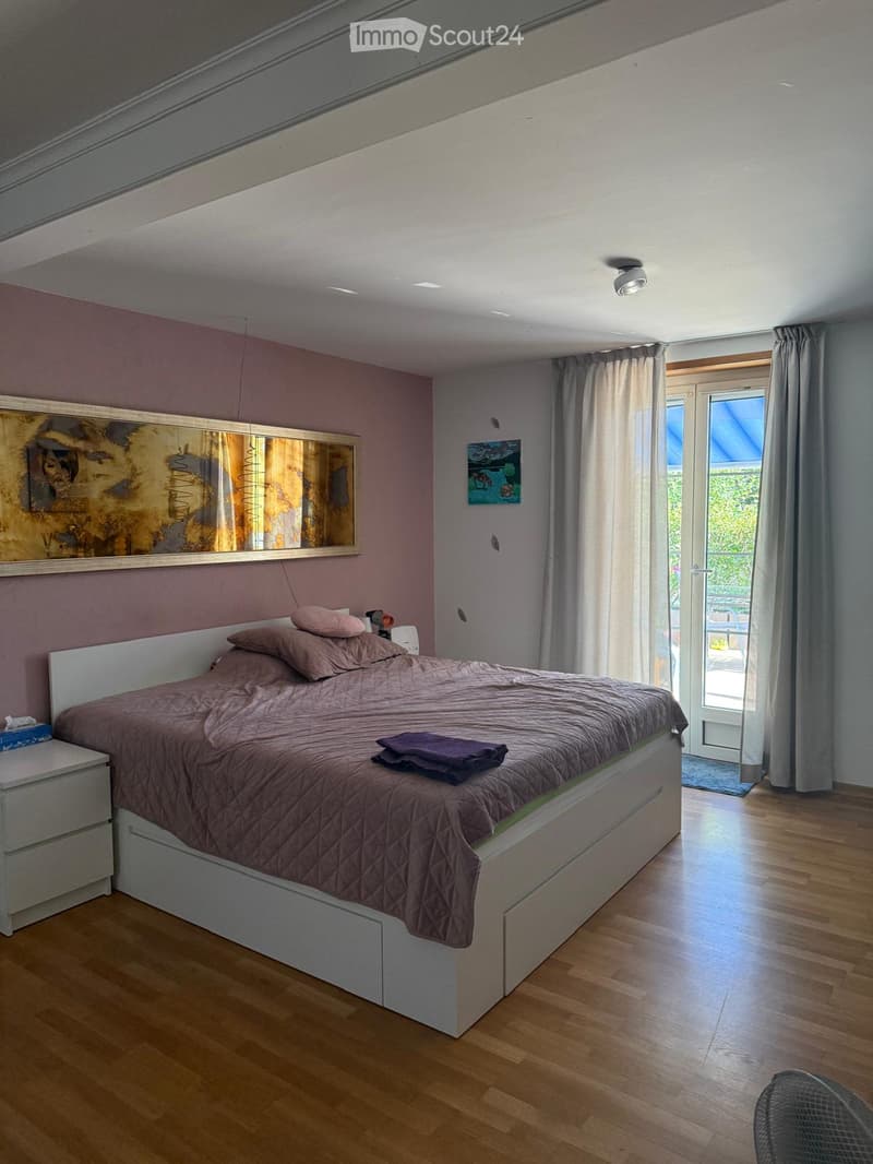 Modern bedroom with a large bed, nightstand, mirror, and artwork on the walls. The room features wooden flooring and large windows with curtains.