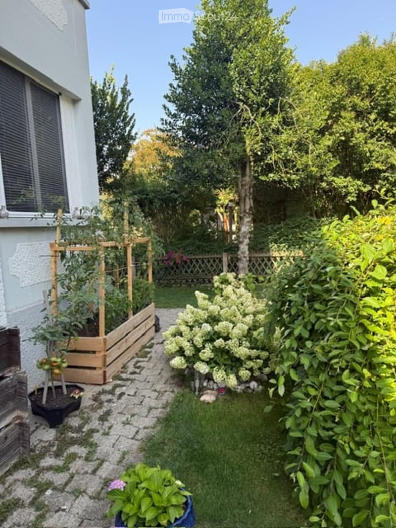 A small, well-maintained garden area with a wooden raised bed, potted plants, a flowering bush, and a paved pathway.