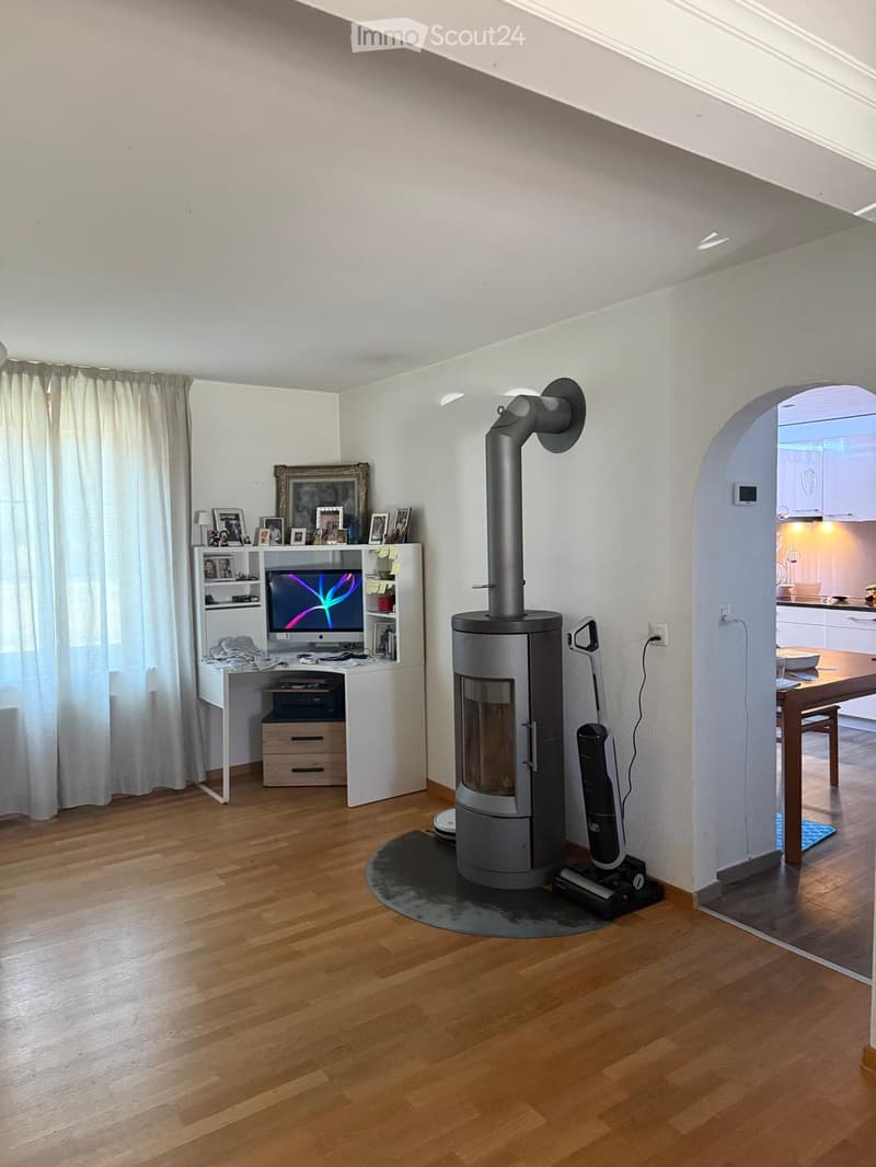 Modern living room with a wood stove, desk with computer, and a view into the kitchen area.
