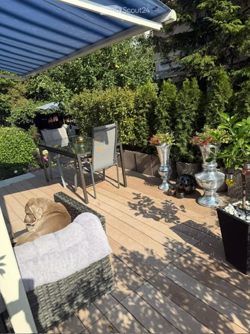 A wooden deck with a covered awning, featuring a wicker sofa, glass table, chairs, and decorative vases with flowers.