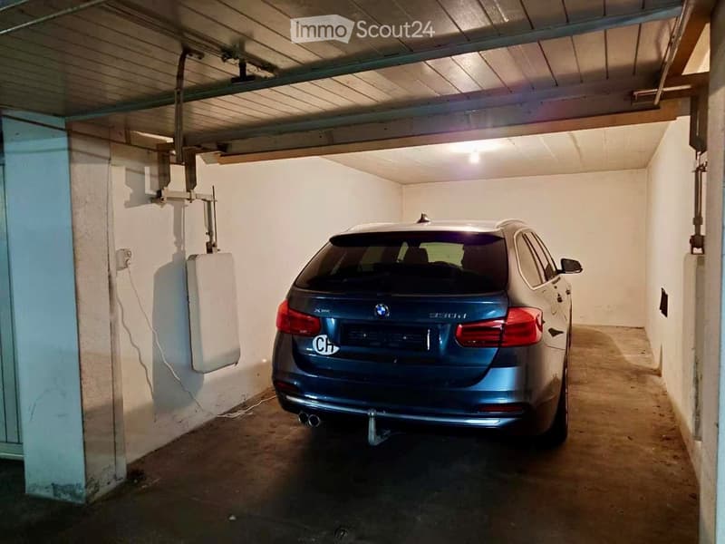 Underground garage with a single parked car, concrete floor, white walls, and a charging station on the left side.